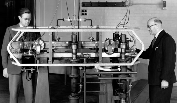 A black-and-white photograph of a man in a suit standing beside a tall rack of electronic equipment in a 1950s laboratory.
