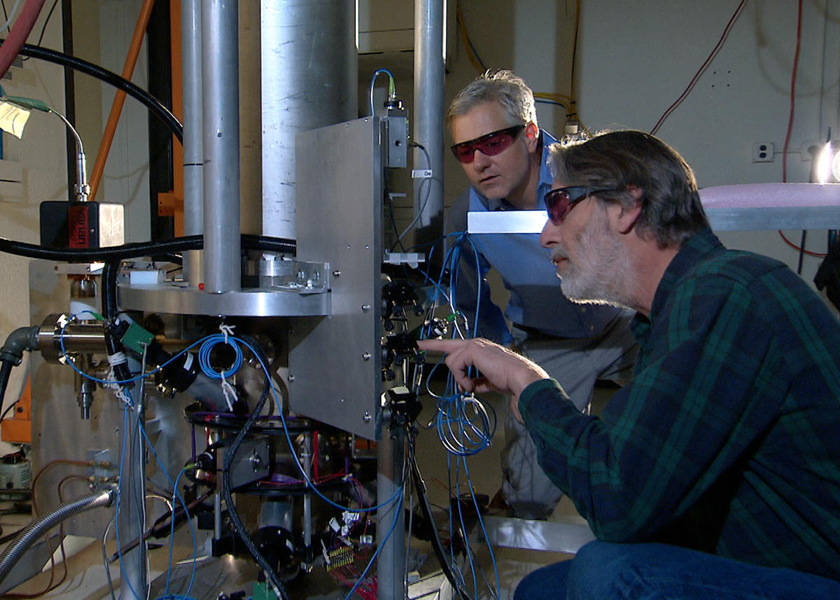 Two NIST physicists standing beside the NIST-F2 caesium fountain atomic clock, a tall cylindrical apparatus wrapped in cabling in a laboratory.